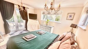 Un dormitorio con edredón verde acolchado, varios almohadones, una araña, televisor de pantalla plana, cómoda y una ventana con cortinas.