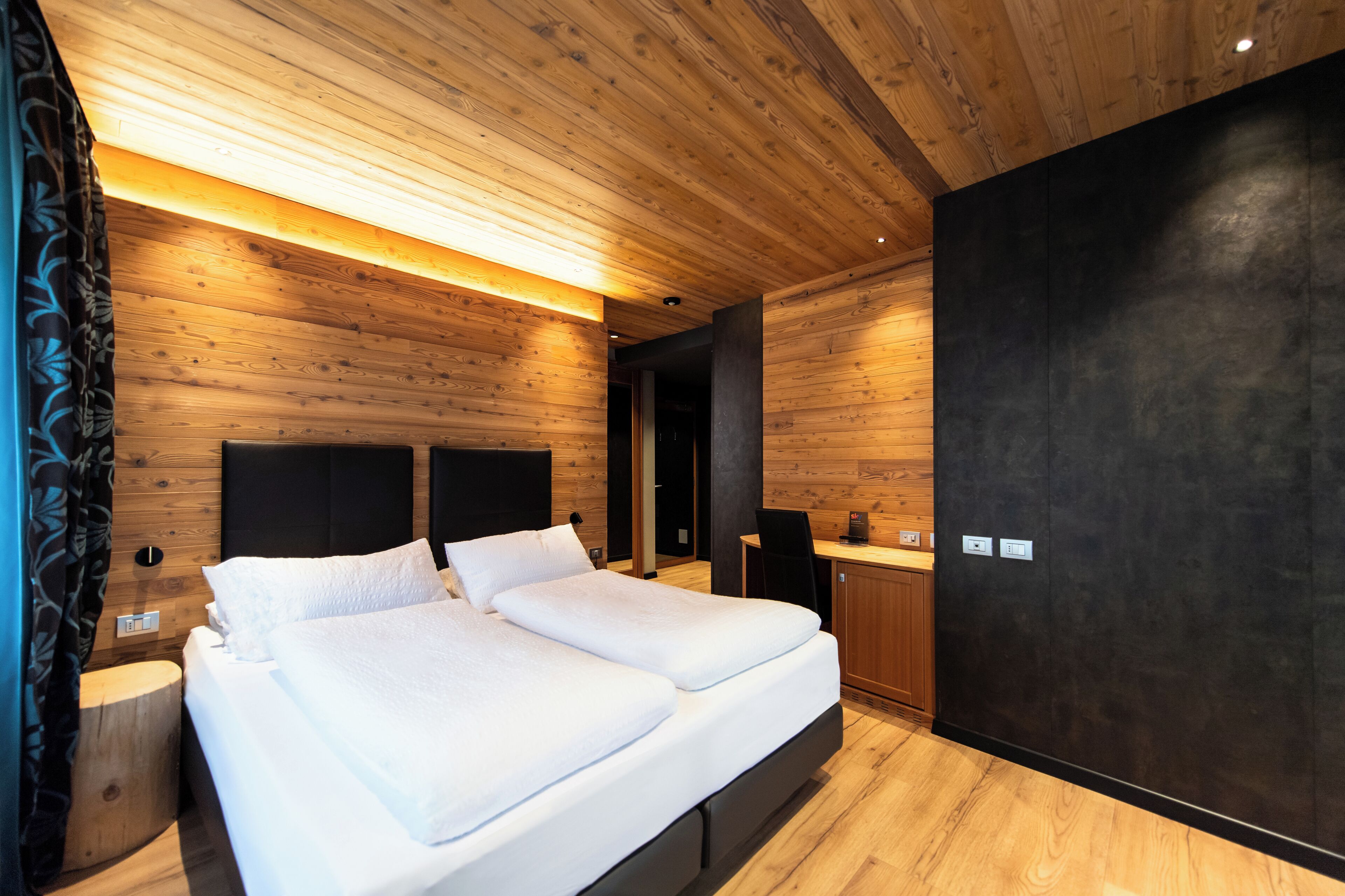 A modern bedroom with a wooden ceiling, a large bed with white bedding, and a dark accent wall.