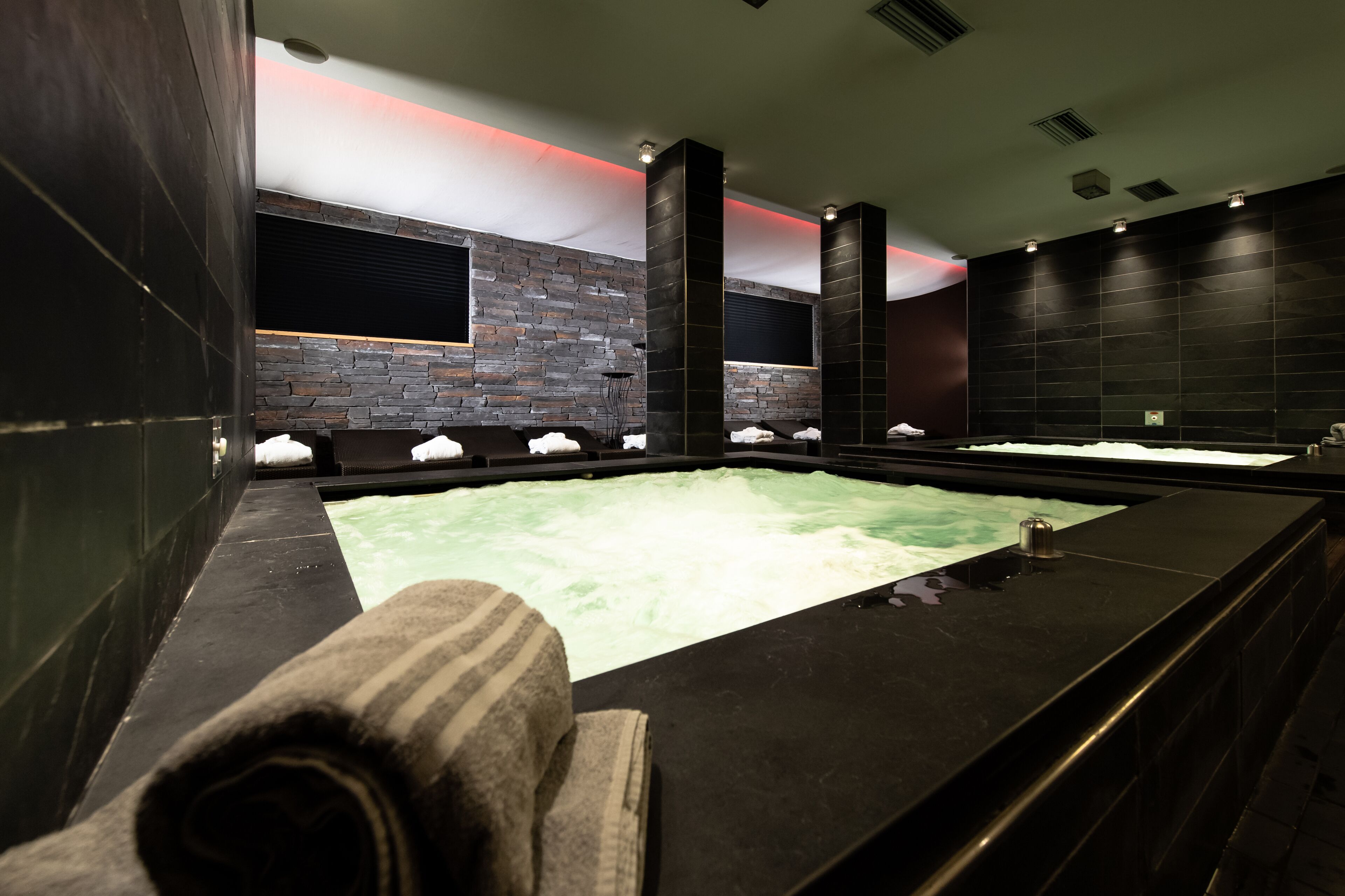 A modern spa with a large hot tub, dark stone walls, and a minimalist design.