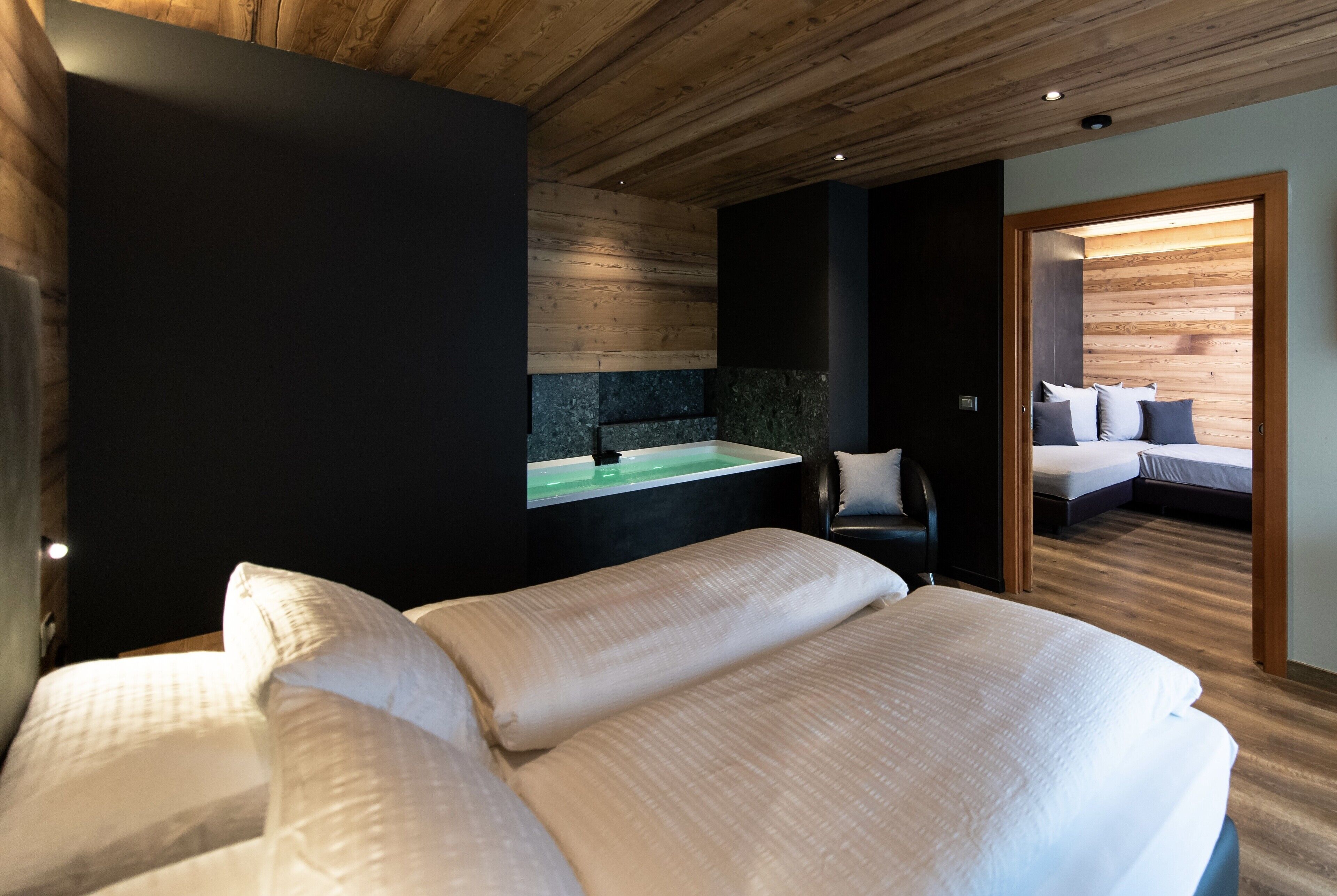 A modern hotel room with a large bed, a small desk with a green sink, and a separate sleeping area with a sofa and pillows.