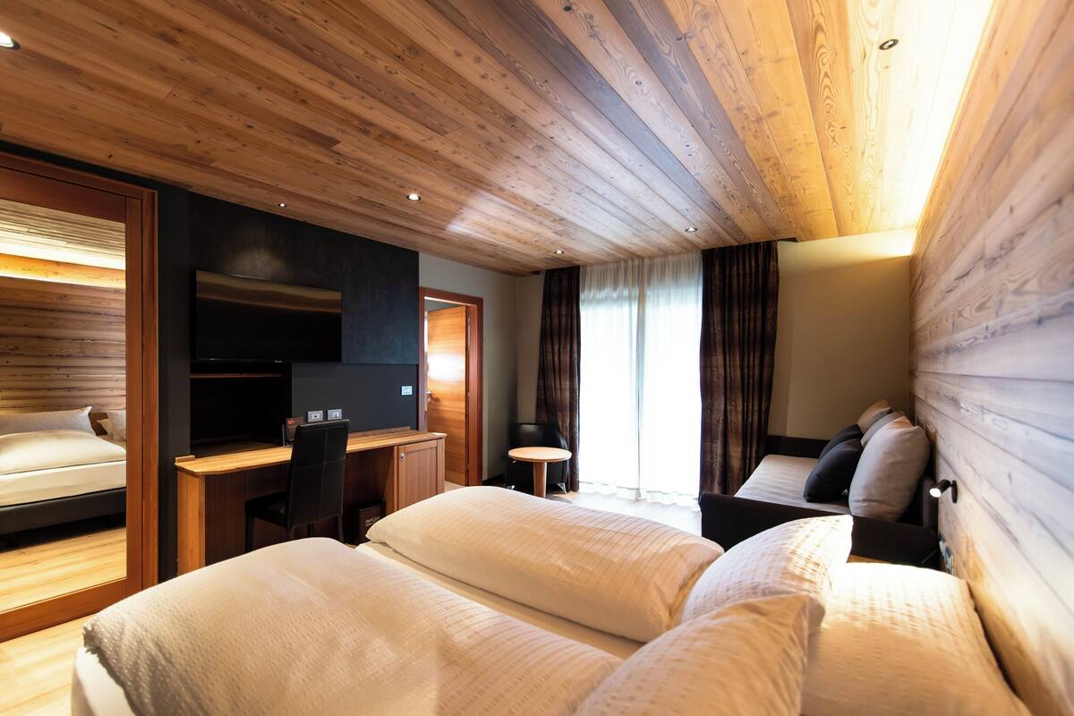 A hotel room with a wooden ceiling, a bed with white bedding, a desk with a chair, a television, and a small table.