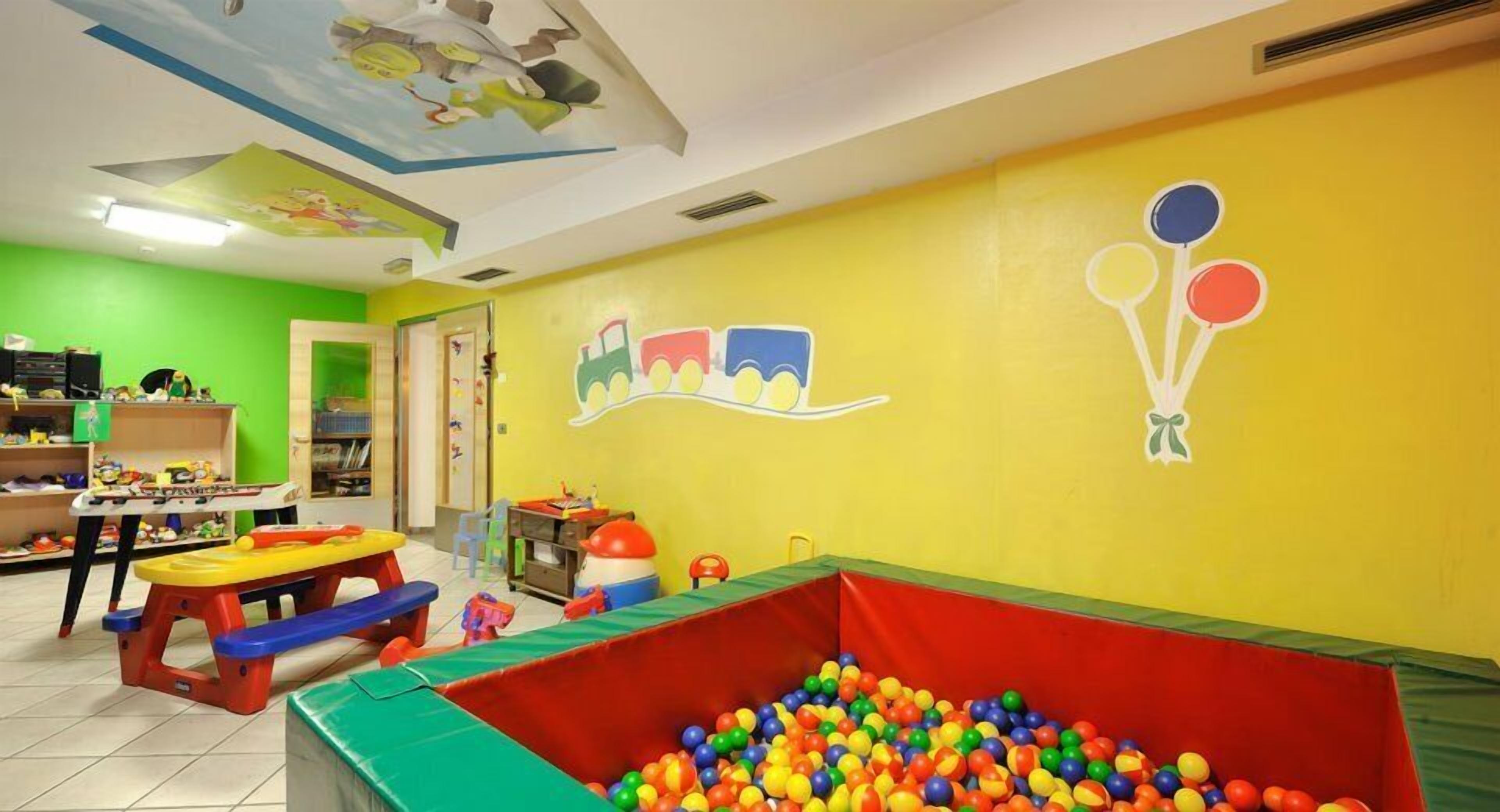 Espaço infantil com piscina de bolinhas, brinquedos coloridos e um mural de um trem e balões na parede.