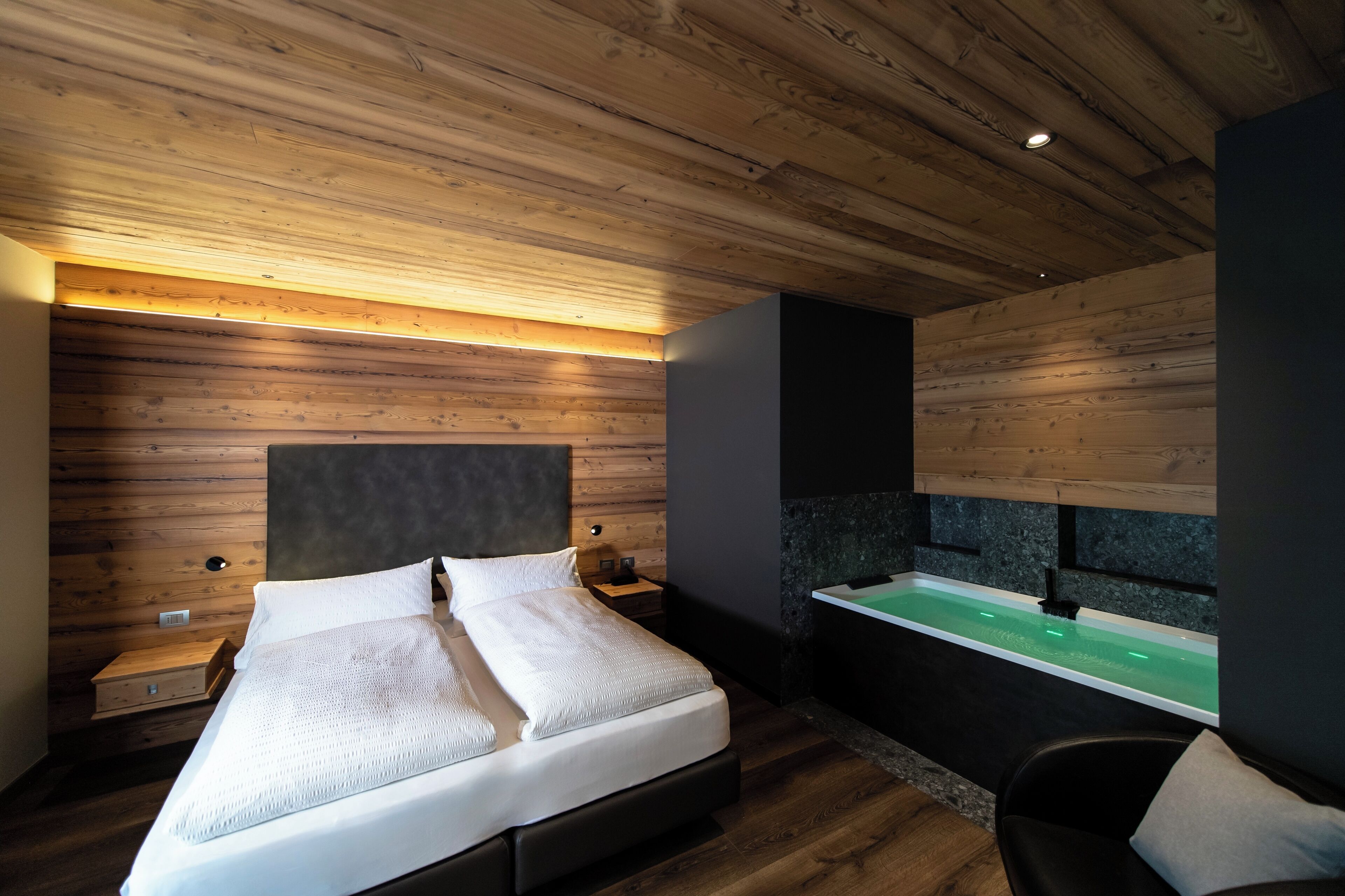 Un dormitorio moderno con techo de madera, una cama grande, una mesita de noche, un televisor montado en la pared y una bañera verde.