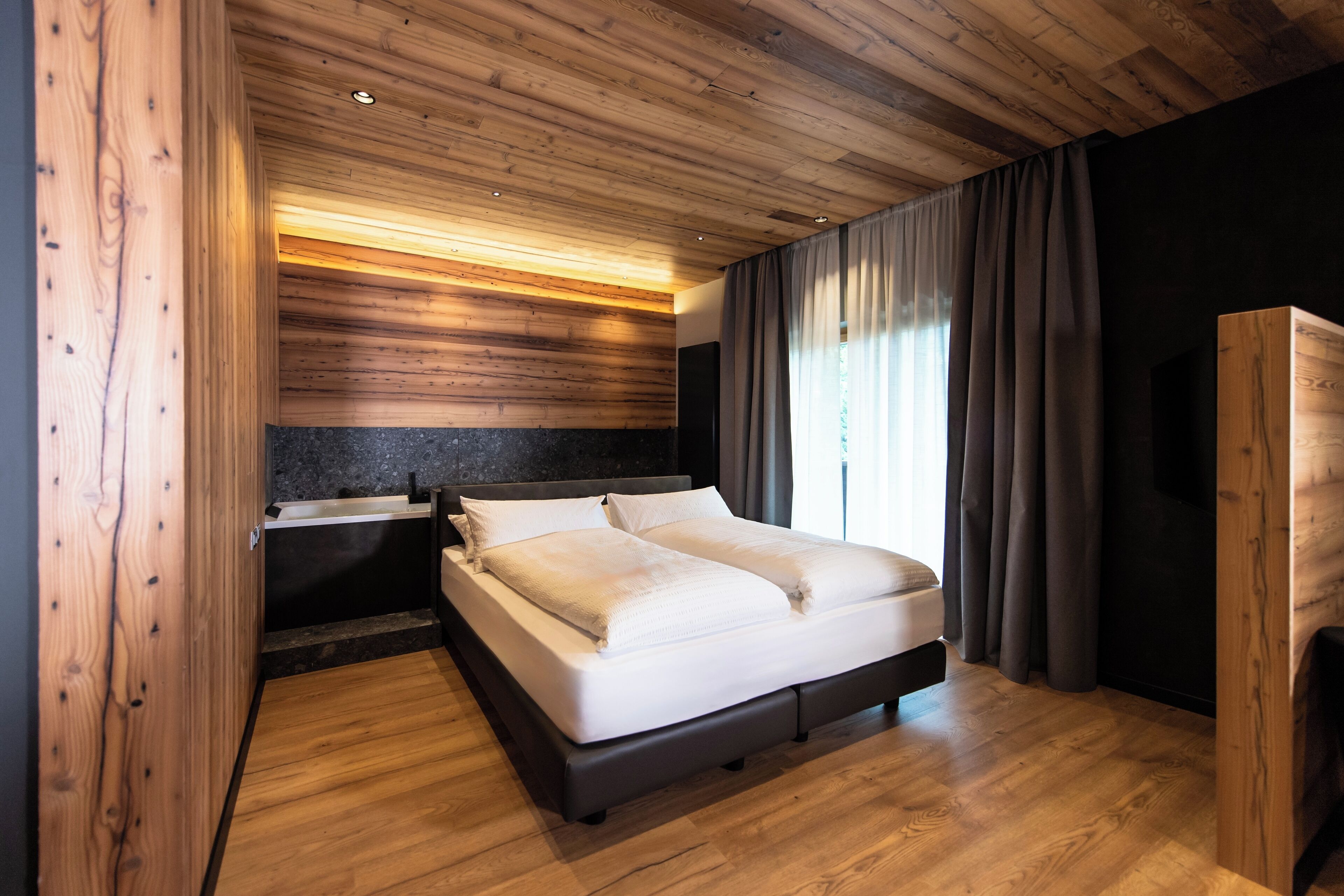 A modern bedroom with a wooden ceiling, a large bed, a dark headboard, a bedside table, and a window with curtains.