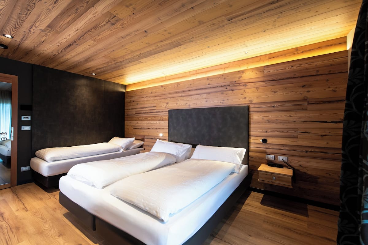 A modern bedroom with wooden panel walls, a large bed, two smaller beds, and a bedside table with a lamp.