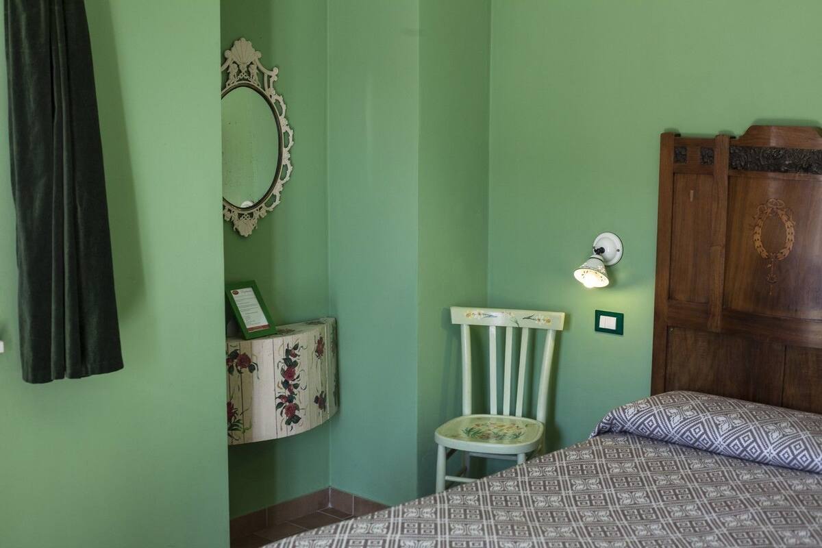A room with a bed, a chair, a wooden wardrobe, a wall-mounted mirror, and a small table with a floral pattern.