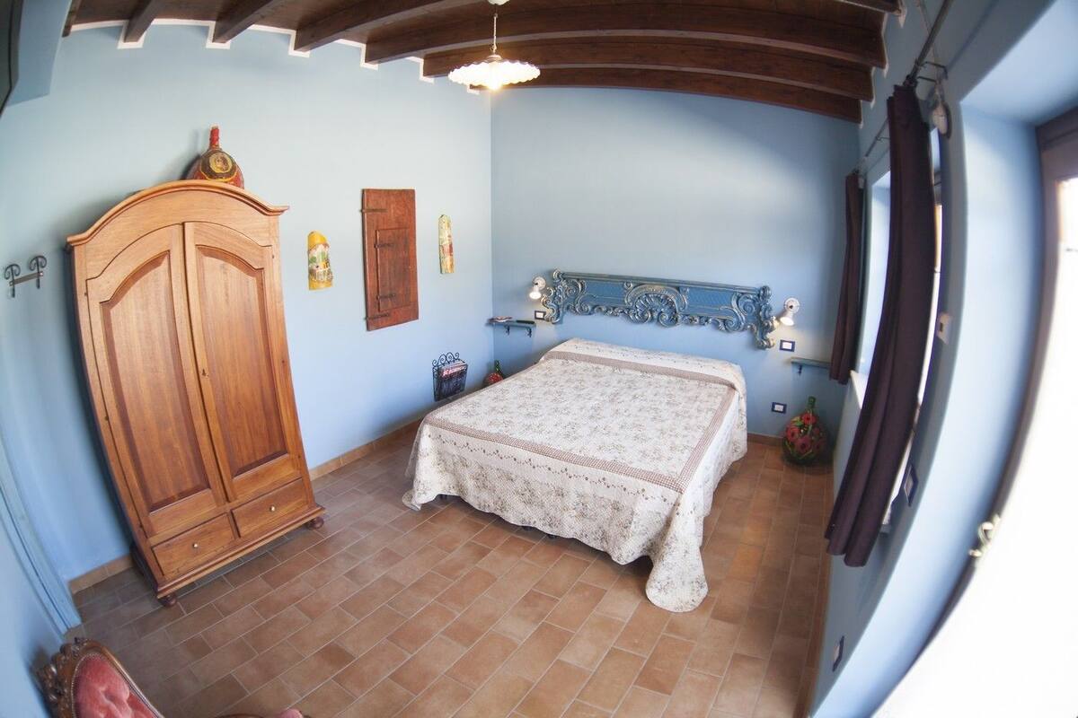 A bedroom with a wooden wardrobe, a bed with a patterned bedspread, a blue headboard, and a tiled floor.