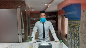 Une personne vêtue d’une chemise blanche et d’une cravate, debout dans une cuisine, avec un plan de travail, un réfrigérateur et une étagère remplie de vaisselle.