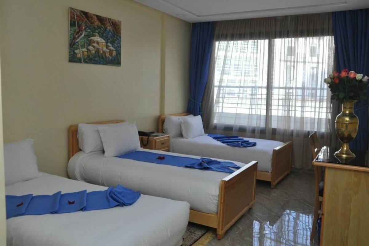 A hotel room with two single beds, a desk, a chair, a vase with flowers, and a painting on the wall.