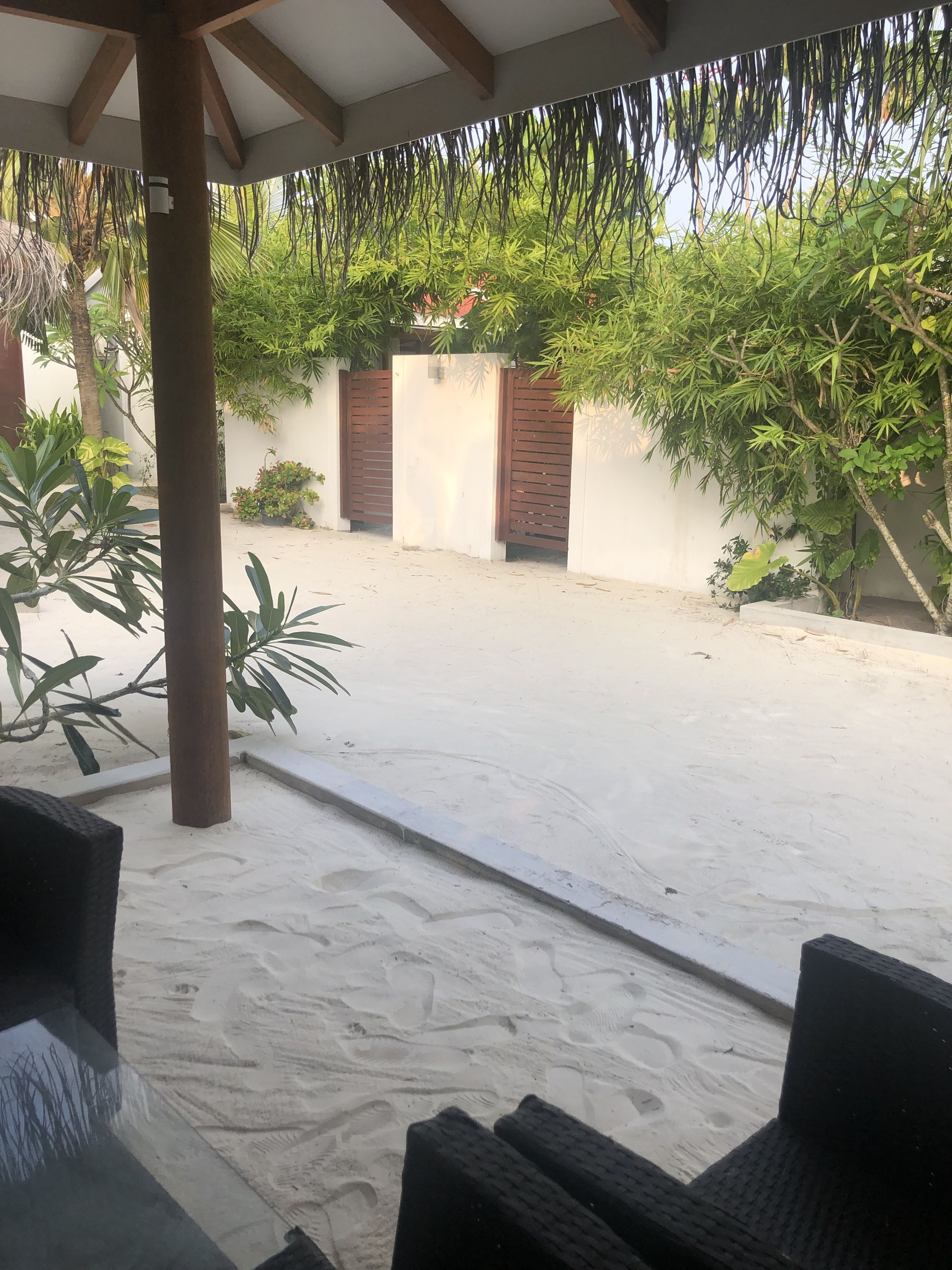 Un espacio al aire libre techado, con suelo de arena, pilares de madera y vistas a un jardín con diversas plantas y una reja.