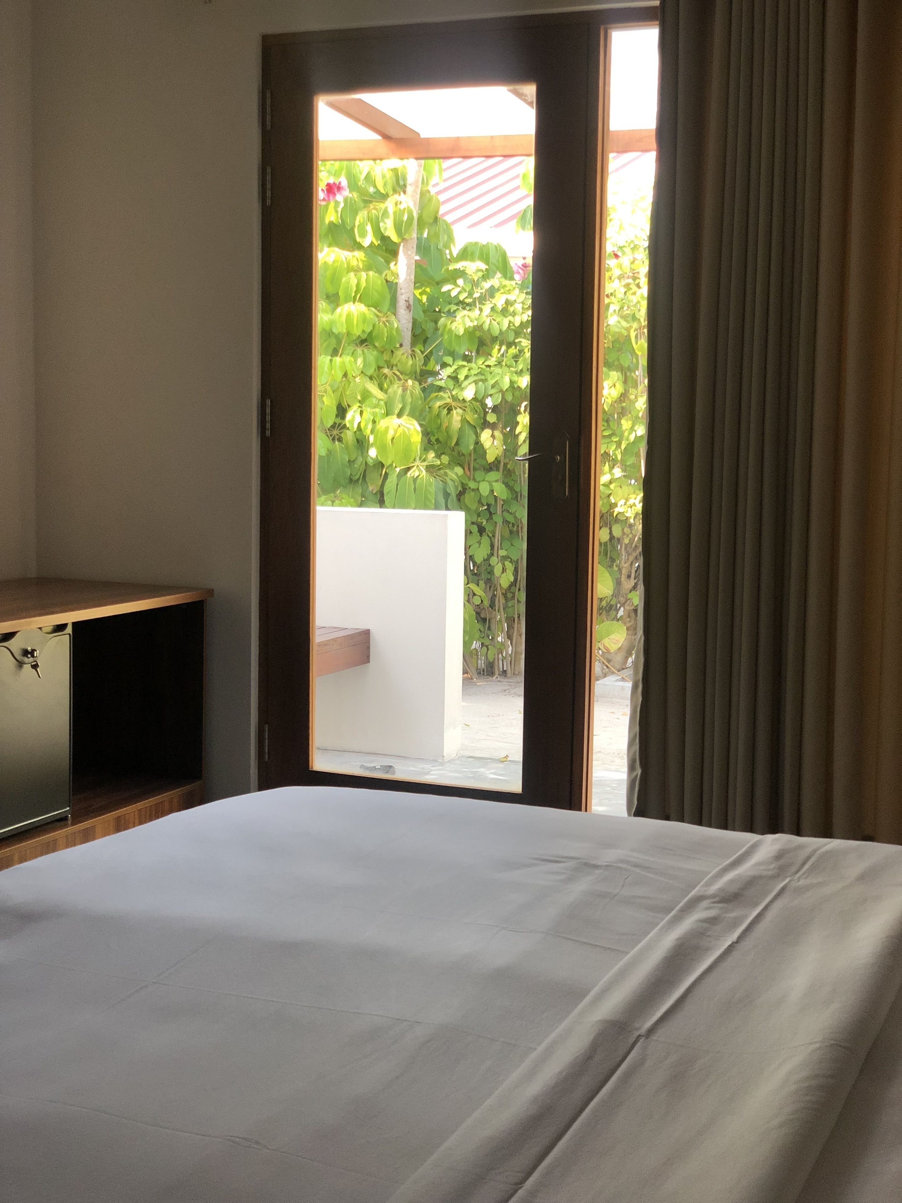 Habitación de hotel con cama, un pequeño mueble de madera y puerta corrediza de vidrio que da a un balcón con follaje.
