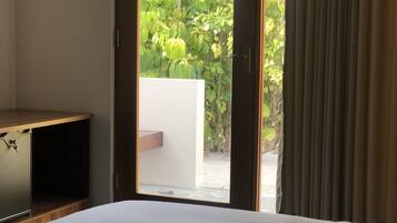 Habitación de hotel con cama, un pequeño mueble de madera y puerta corrediza de vidrio que da a un balcón con follaje.