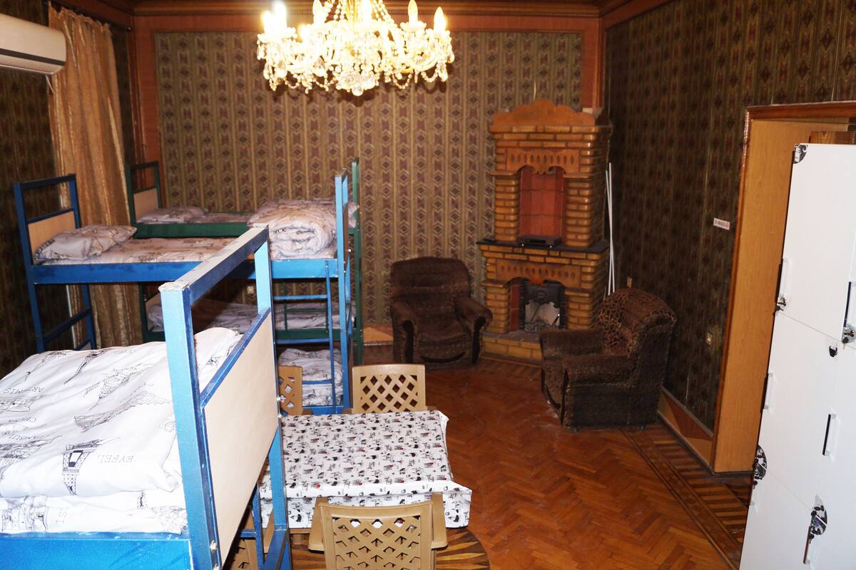 Ein Zimmer mit Etagenbetten, einem kleinen Tisch, zwei Stühlen, einer Kandelaber und einem Kamin, sowie einem Kleiderschrank.
