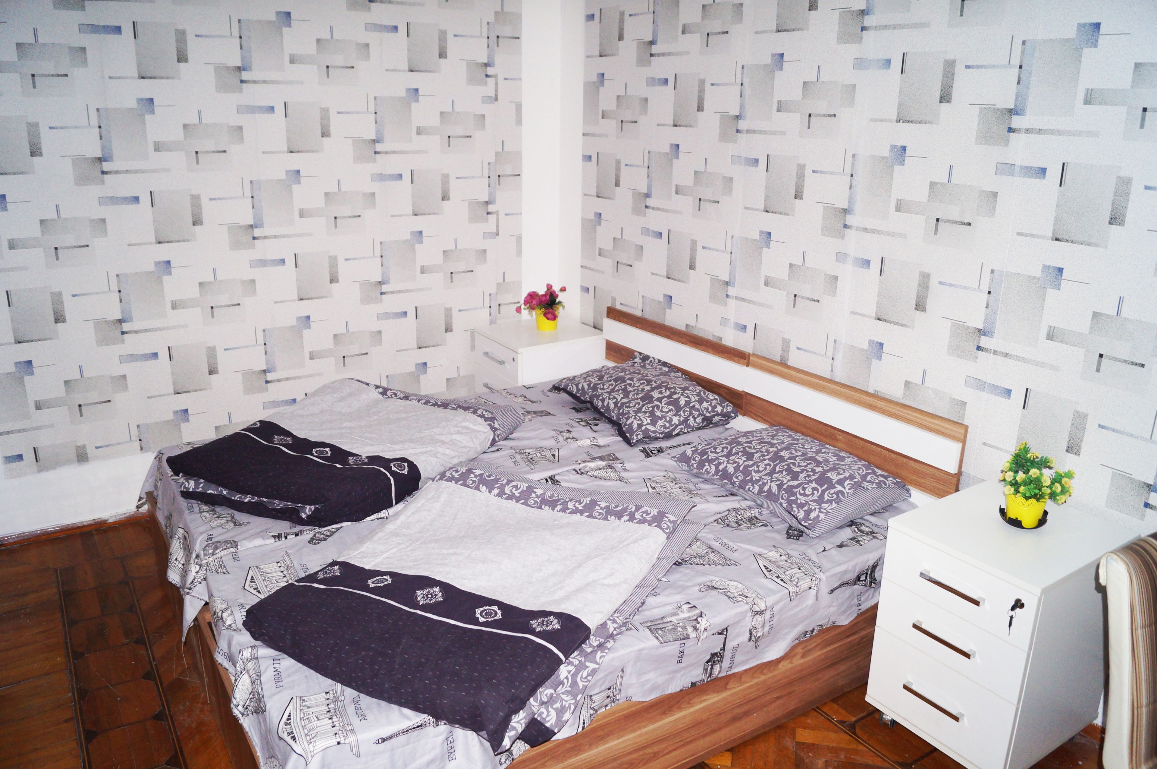 A neatly made bed with patterned bedding, a bedside table with a vase of flowers, and a white cabinet.