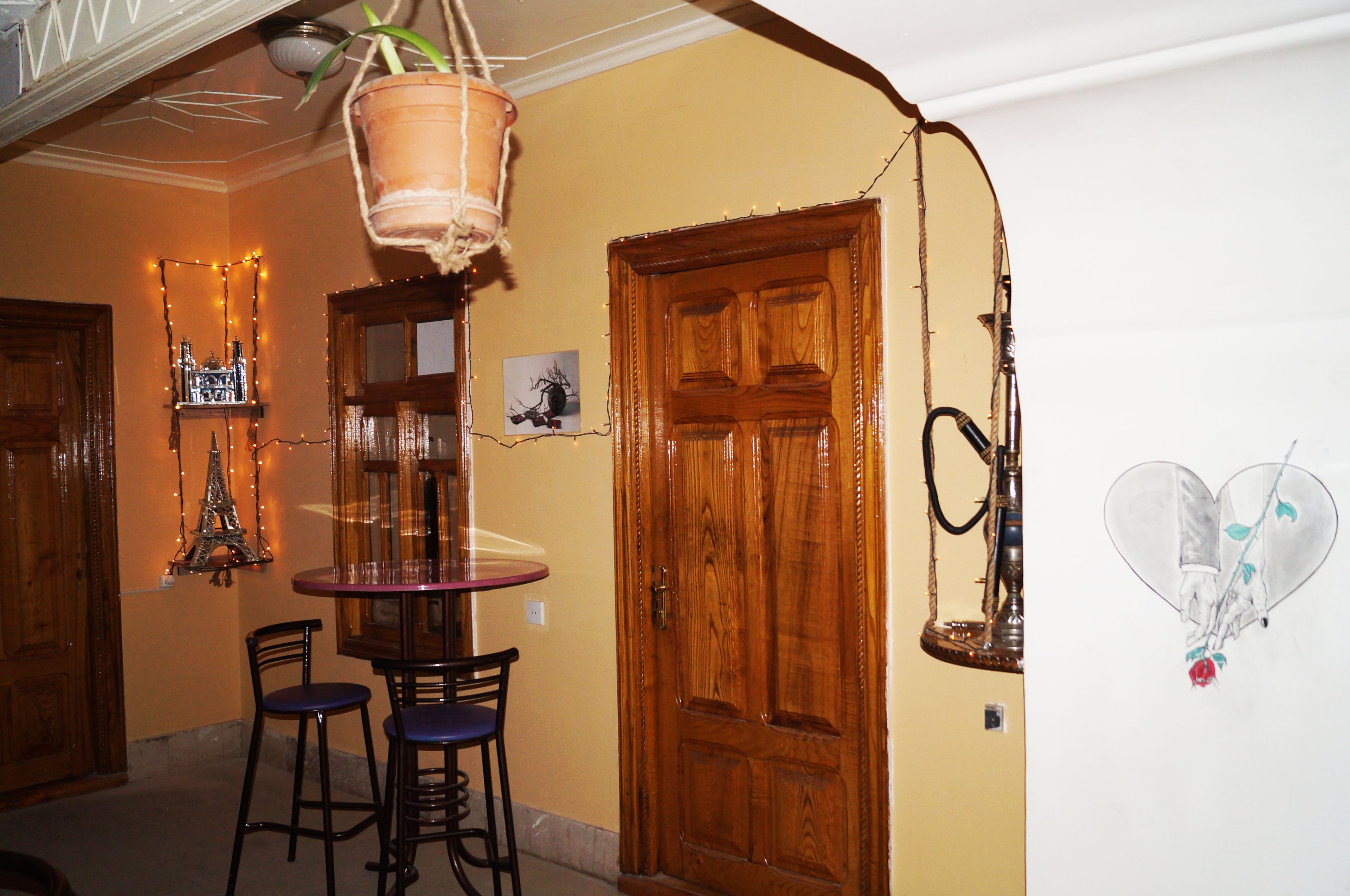 Une pièce avec une porte en bois, une plante suspendue, une table avec des chaises et un mur orné d’un tableau et de guirlandes lumineuses.