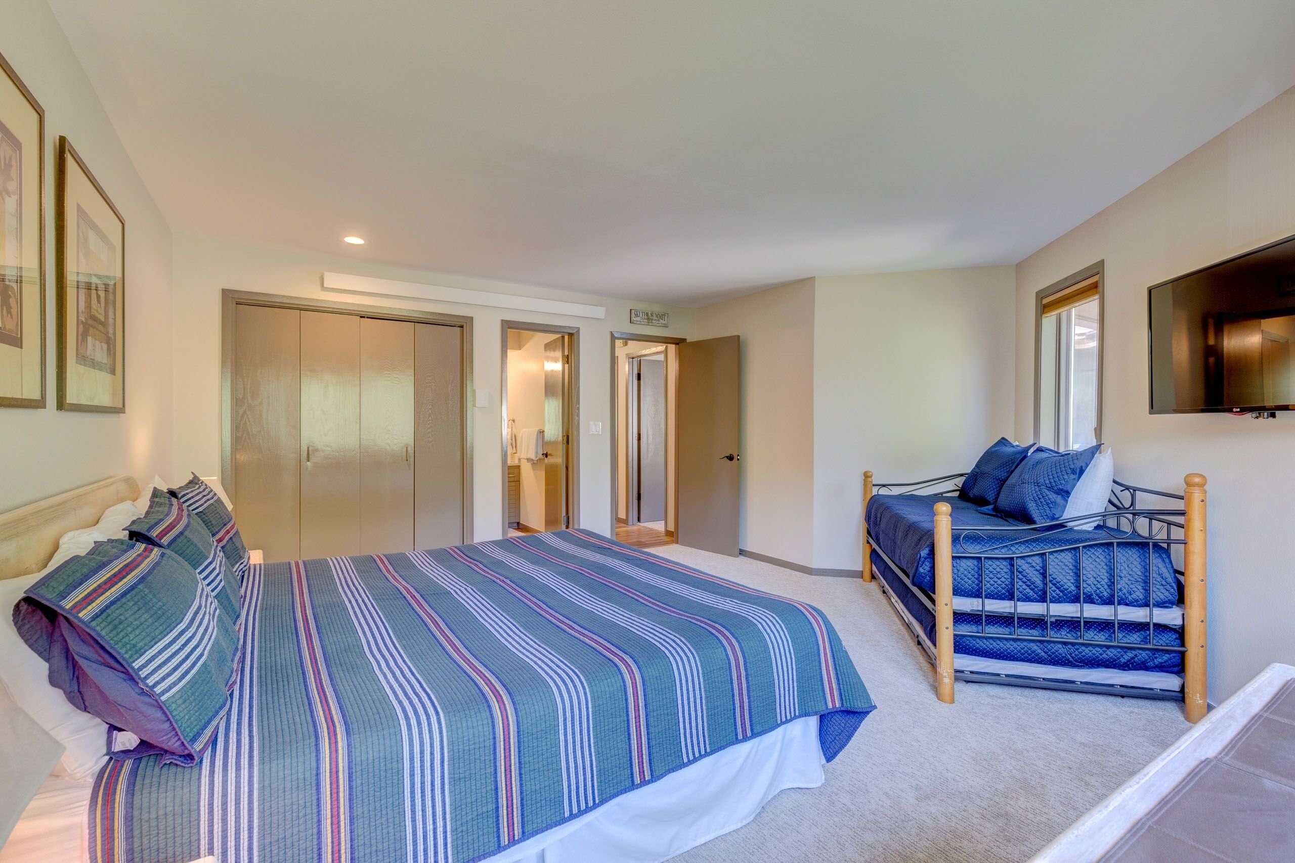 Un dormitorio con una cama grande, una cama más pequeña y un armario. Hay un televisor en la pared y una puerta que da a otra habitación.