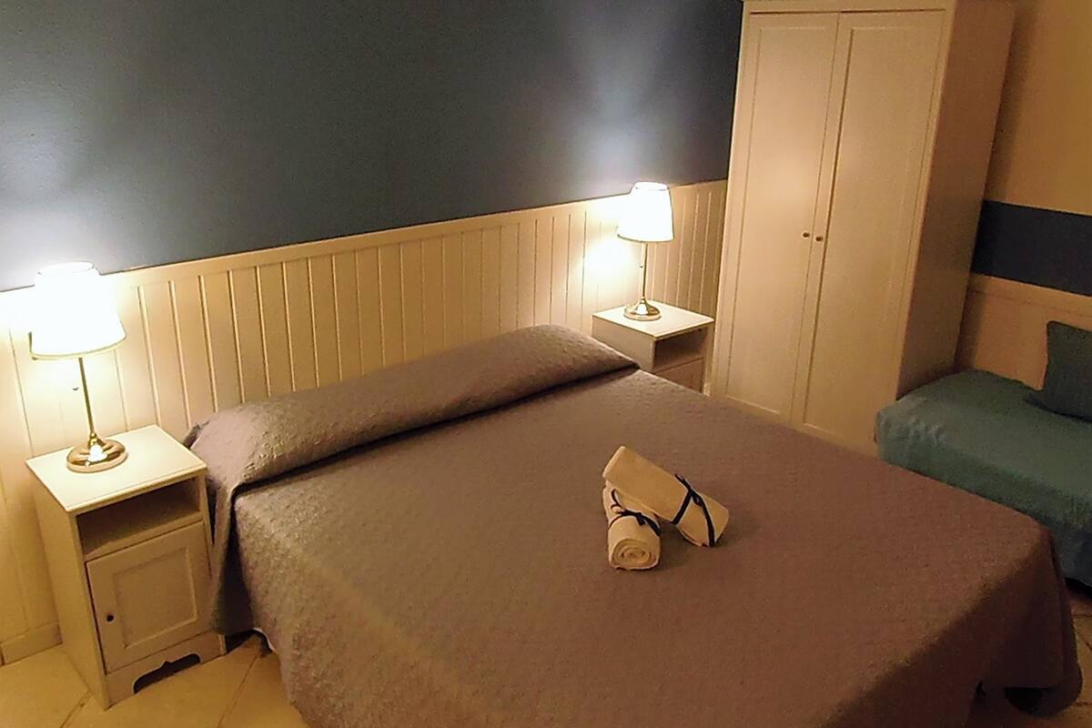 Een opgemaakt bed met een bruine spreideken en een opgerolde handdoek, twee bedlampen, een nachtkastje, een garderobe en een groene kussen op een blauw bed in een hotelkamer.