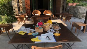 Area pranzo all'aperto con sedie in vimini, un tavolo apparecchiato con piatti, tazze e diversi cibi.