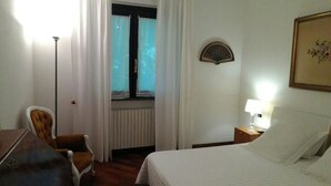 Camera d'albergo con un letto, una sedia, una scrivania, una lampada, una finestra con tende e un ventilatore a parete.