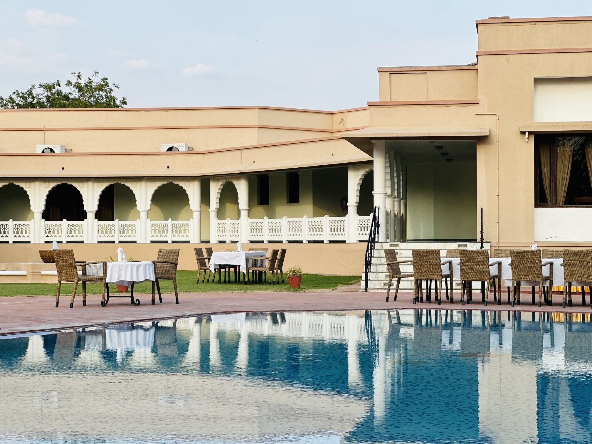 Zona de piscina con mesas y sillas, una piscina e edificio con arcos y balcón.