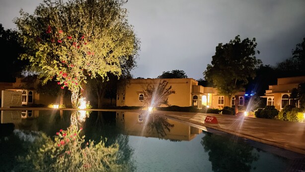 Vue nocturne d’un hôtel avec une piscine qui reflète les arbres et les bâtiments environnants.