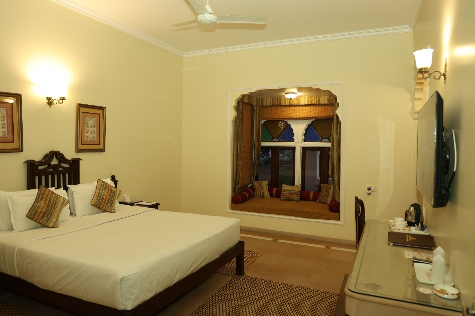 Quarto de hotel com cama, escrivaninha com espelho, televisão e janela com vista exterior.