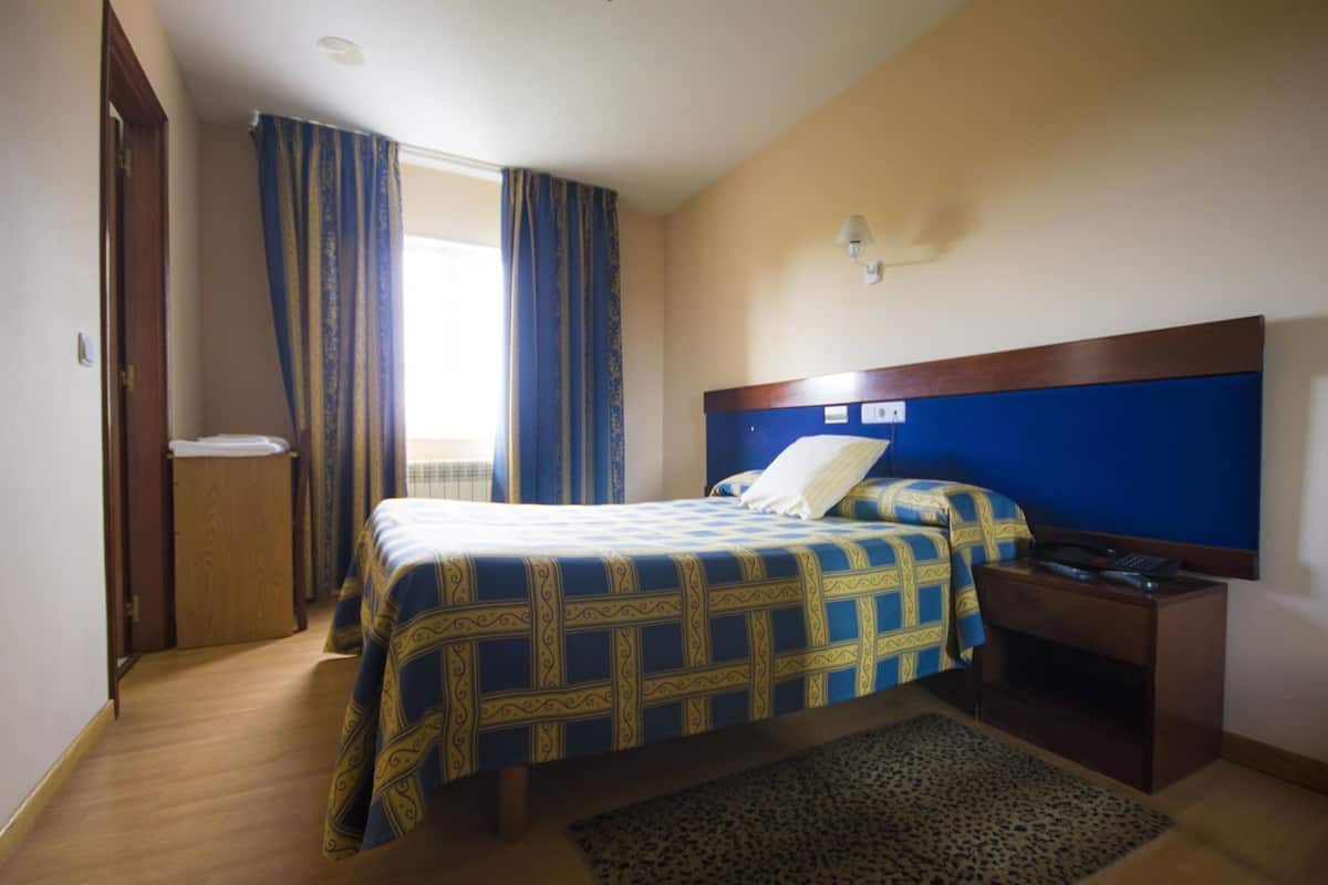 A hotel room with a bed, bedside table, lamp, and a view of the outdoors.