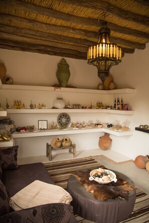 Una sala de estar acogedora con un pouf redondo, un sofá y una lámpara colgante. Estantes exhiben diversos objetos decorativos y botellas.