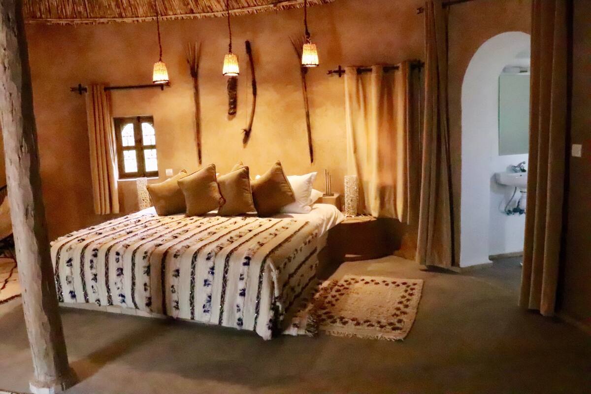 Quarto com cama, travesseiros, mesa de cabeceira, abajur, janela e cortinas.