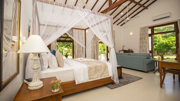 A bedroom with a canopy bed, a bedside table with a lamp, a small table with a vase of flowers, a sofa, and a dining table.