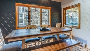 Un espace repas avec une table en bois et des bancs rembourrés bleus, de grandes fenêtres à cadre en bois et un luminaire suspendu au plafond.