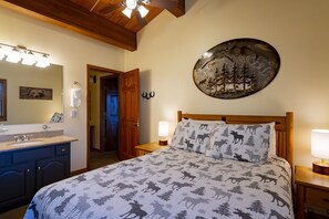 Een slaapkamer met een bed, nachtkastje, bedlamp, houten hoofdbord, een wanddecoratie, een badkamer met wastafel en spiegel, en een deur naar een andere kamer.