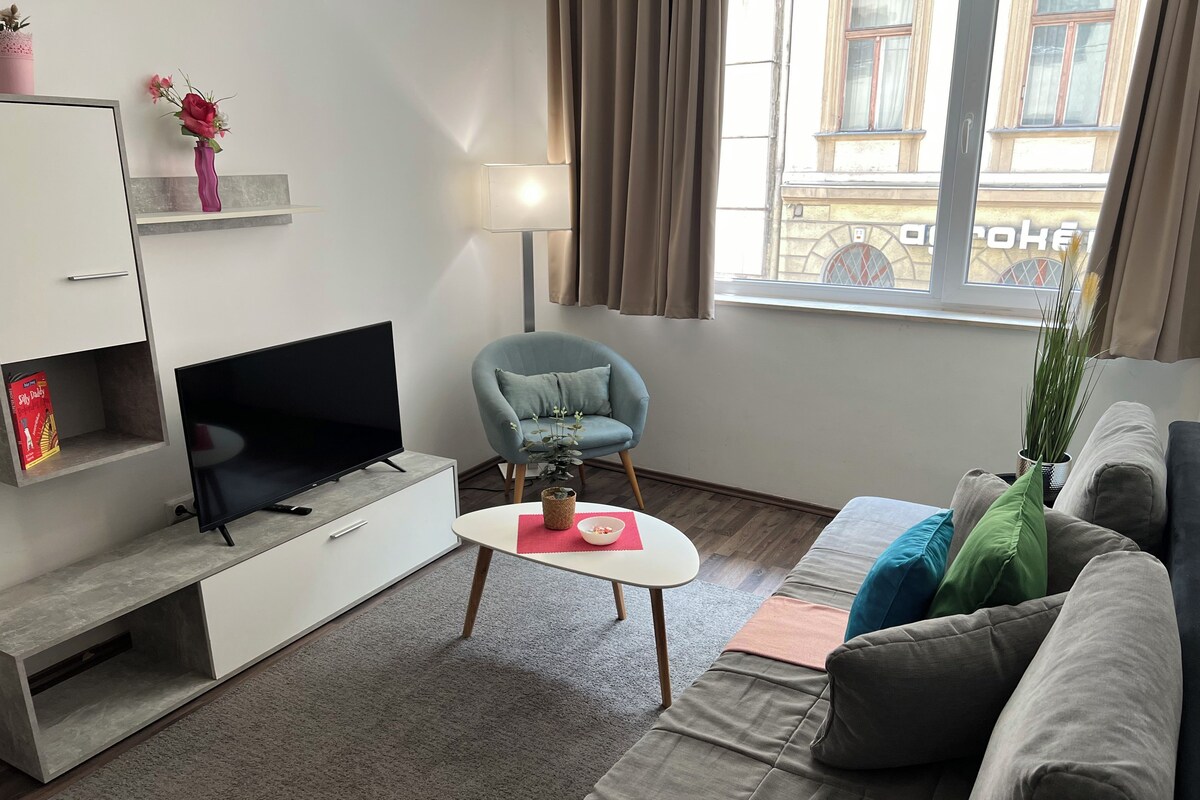 A modern living room with a flat-screen TV, a grey sofa, a blue armchair, and a small table with a vase.