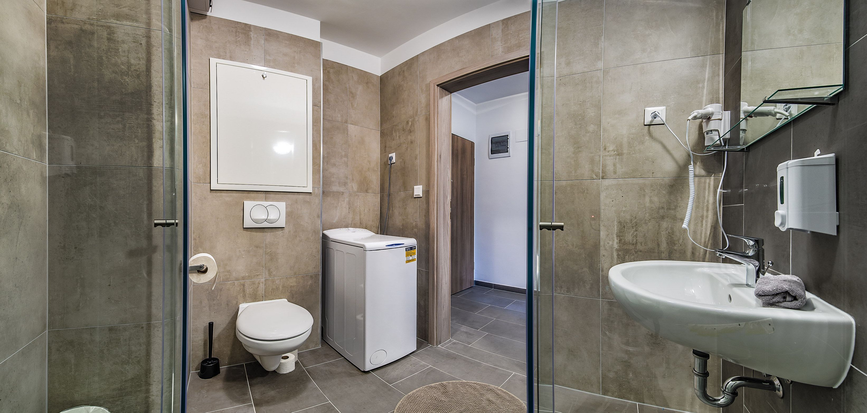 A modern bathroom with a toilet, a compact washing machine, a glass shower enclosure, and a sink with a towel.