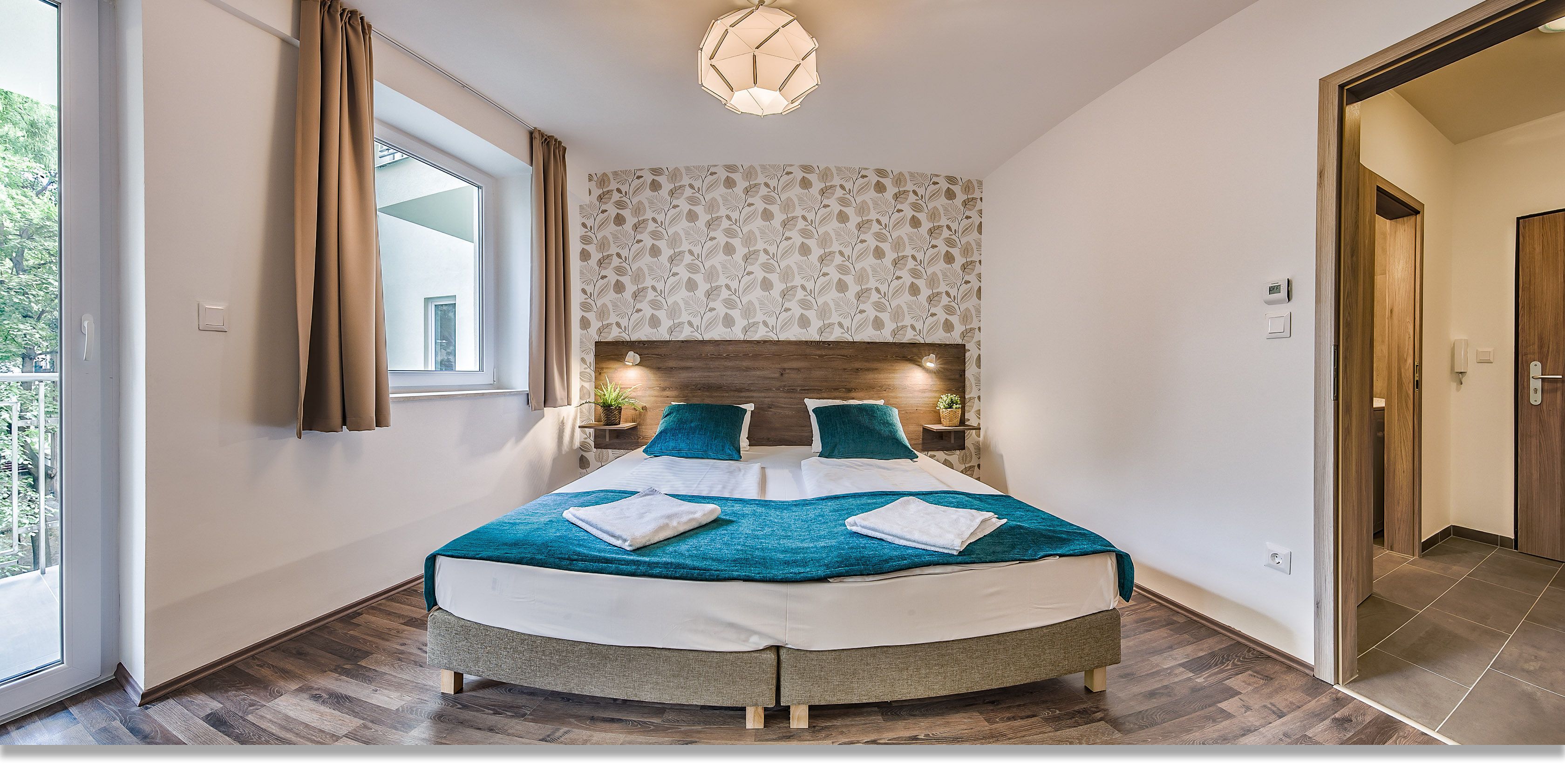 Een moderne hotelkamer met een groot bed, een houten hoofdeinde, uitzicht door een raam en een deur naar een andere kamer.