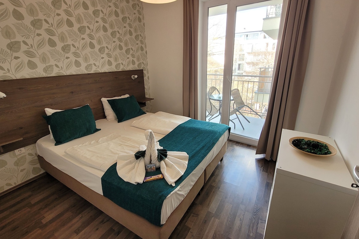 A hotel room with a wooden headboard, a bed with white linens and teal pillows, a small table with a bowl of fruit, and a balcony with outdoor furniture.