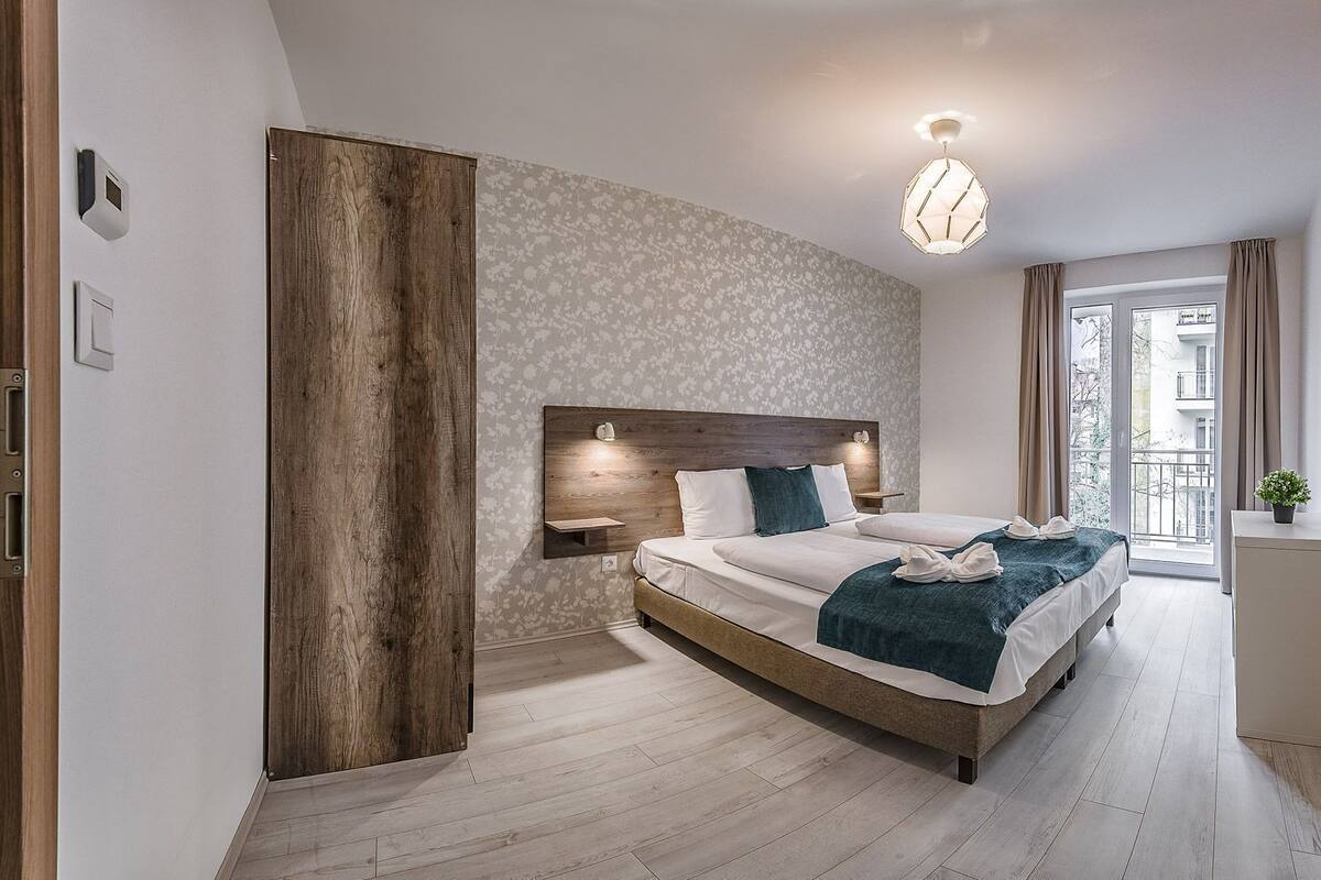 Une chambre moderne avec un grand lit, une tête de lit en bois, une petite table de chevet et une vue sur l’extérieur par une fenêtre.