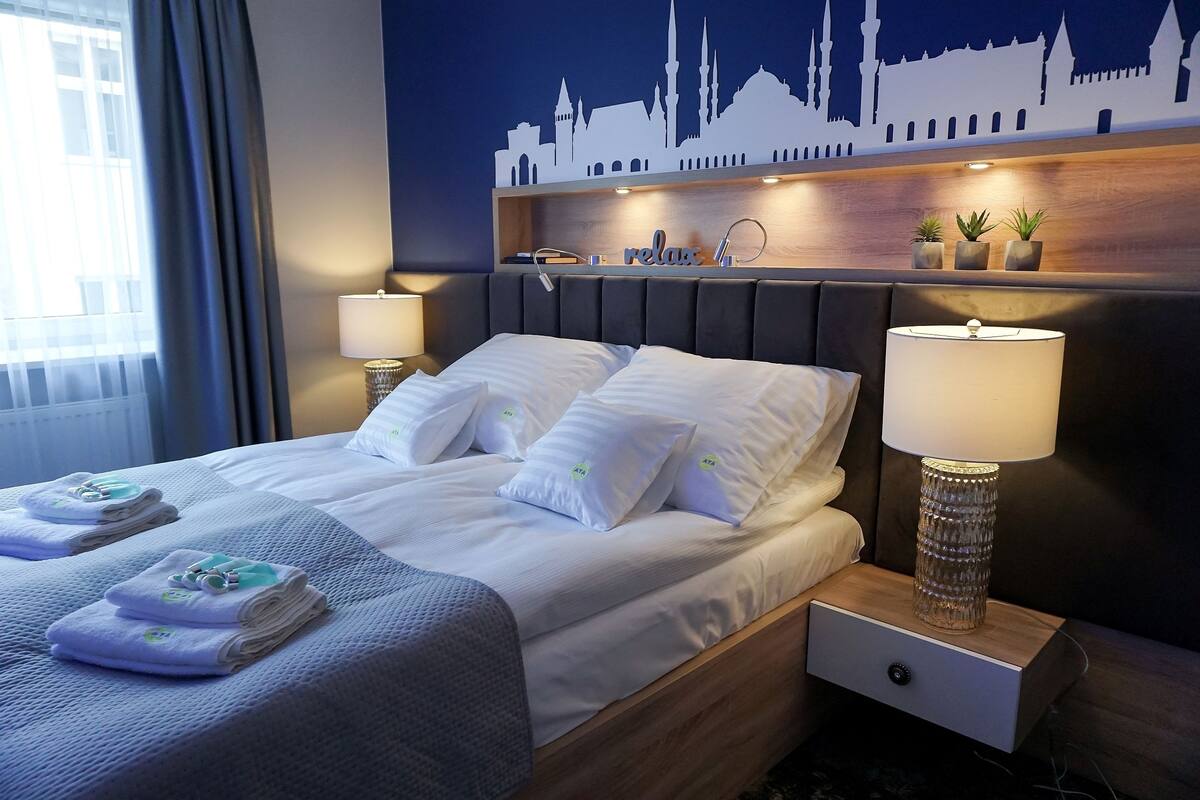 Une chambre d’hôtel comprenant un lit, deux tables de chevet, une petite table, une décoration murale et une fenêtre avec des rideaux.