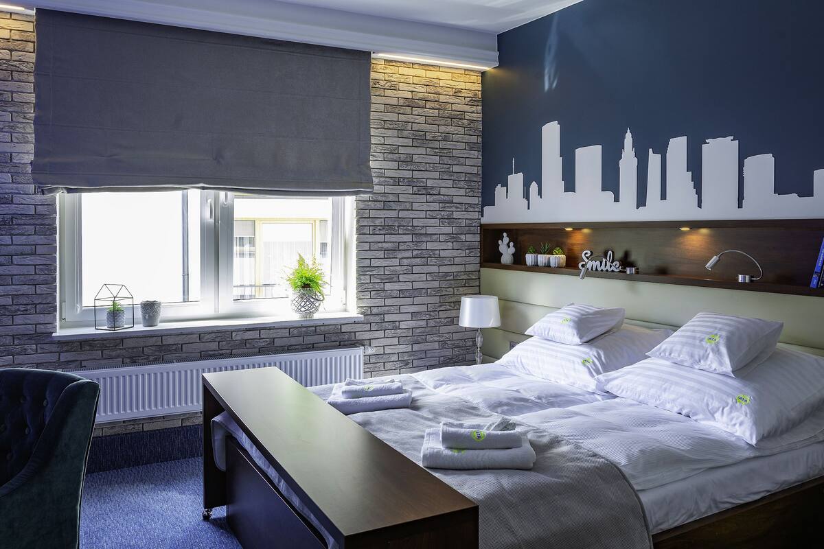 Une chambre d’hôtel comprenant un lit, un bureau, une chaise, une fenêtre avec vue, une fresque représentant le panorama de la ville et un radiateur.