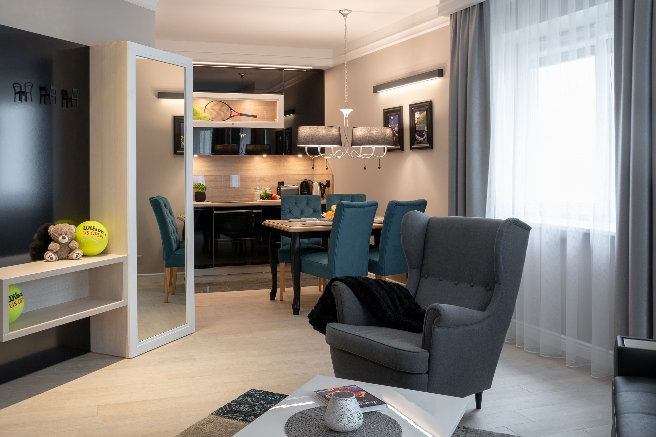 Uma sala de estar moderna com uma poltrona cinza, cadeiras azul-turquesa, uma mesa de centro e uma TV em uma prateleira. Uma cozinha é visível ao fundo, com uma mesa de jantar e cadeiras.