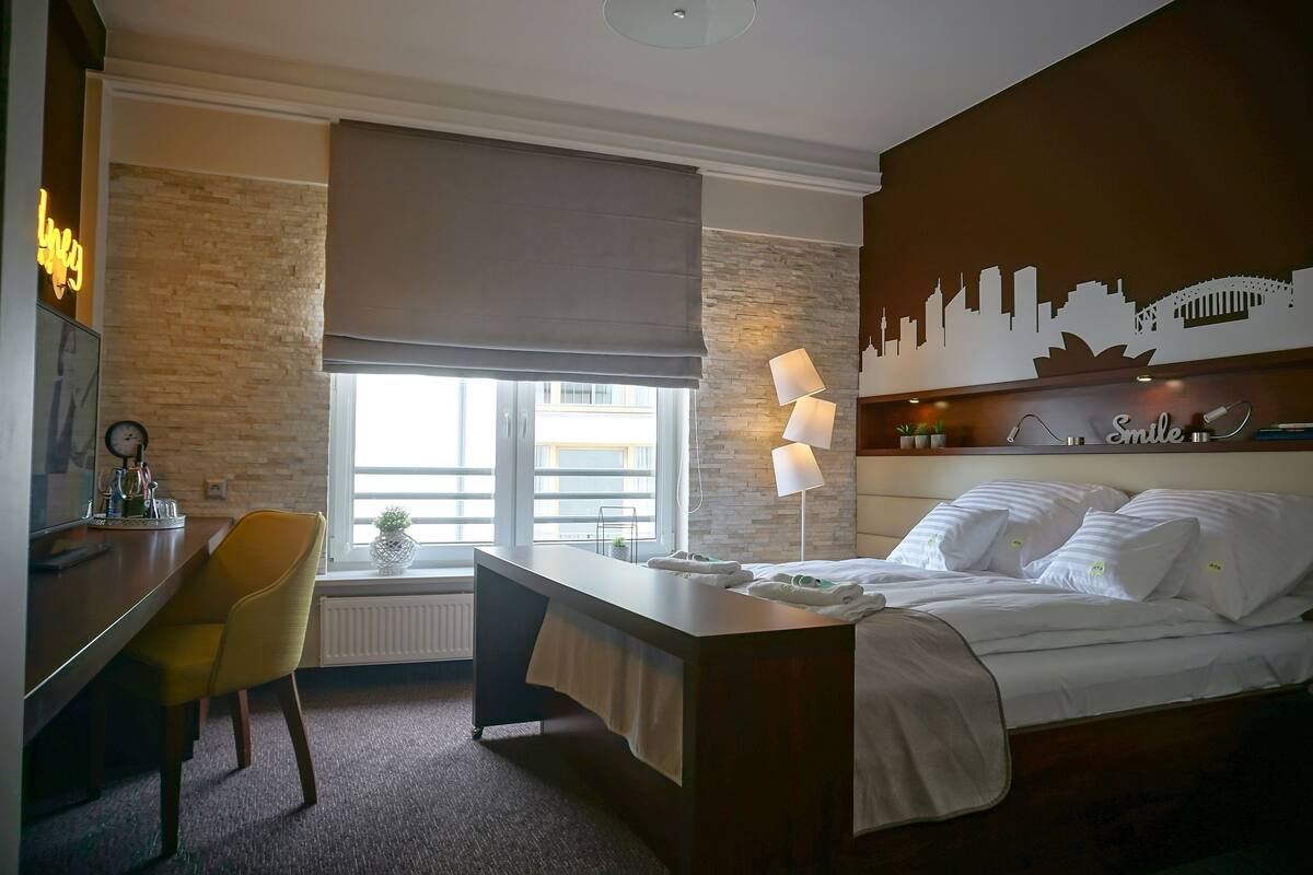 Une chambre d’hôtel avec un lit, un bureau, une chaise, une fenêtre donnant sur un beau paysage et une fresque représentant la vue panoramique de la ville sur un des murs.
