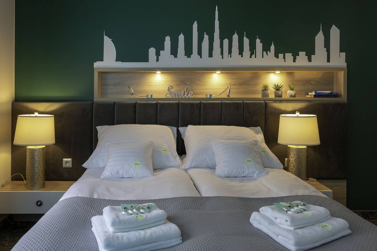 Une chambre d’hôtel avec une silhouette du paysage urbain peinte sur le mur, un lit avec du linge de maison blanc, deux lampes de chevet et une étagère avec des livres et des plantes.