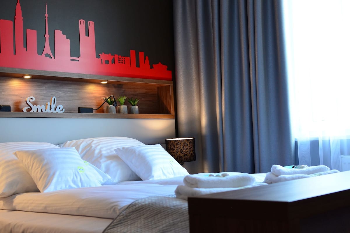 Une chambre d’hôtel avec un lit, des oreillers, une table de chevet avec une lampe, un mur orné d’une silhouette représentant le profil de la ville, et une fenêtre avec des rideaux.