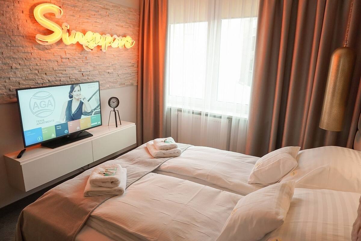 Une chambre d’hôtel avec un mur en briques, un néon, un téléviseur affichant le texte « AGA TENIS APARTMENTS », et deux lits avec des serviettes.