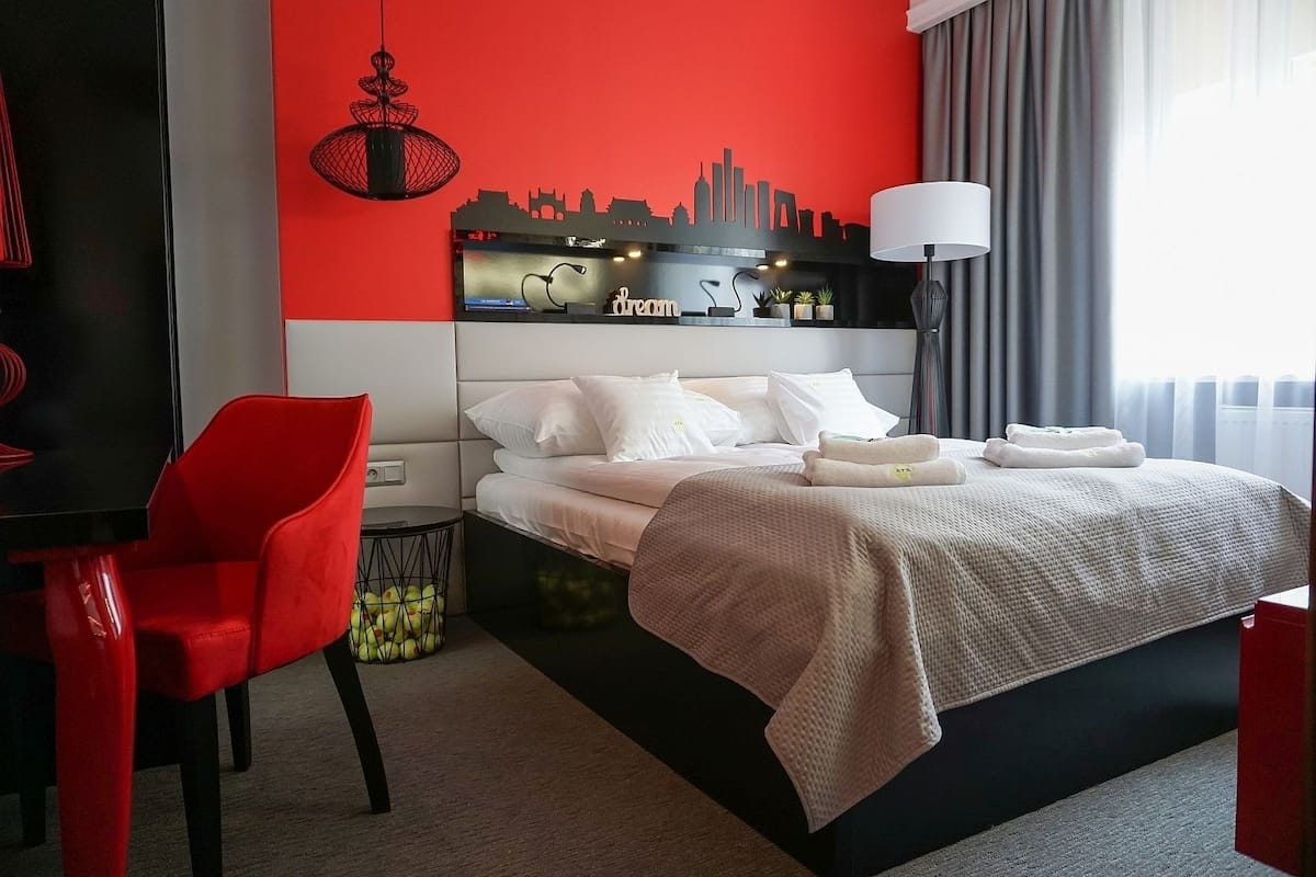Une chambre d’hôtel avec un lit, un fauteuil rouge, une décoration murale représentant une vue panoramique de la ville, une lampe et une fenêtre avec des rideaux.