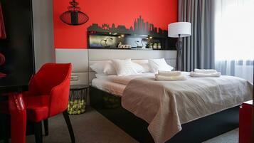 A hotel room with a bed, a red chair, a city skyline wall art, a lamp, and a window with curtains.