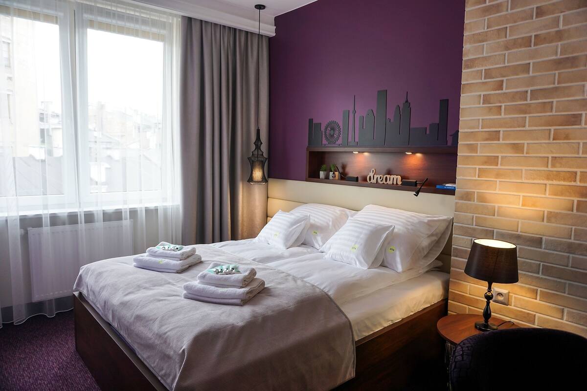 Une chambre d’hôtel avec un lit, une table de chevet avec une lampe, une décoration représentant le panorama de la ville sur le mur, et une fenêtre avec des rideaux.