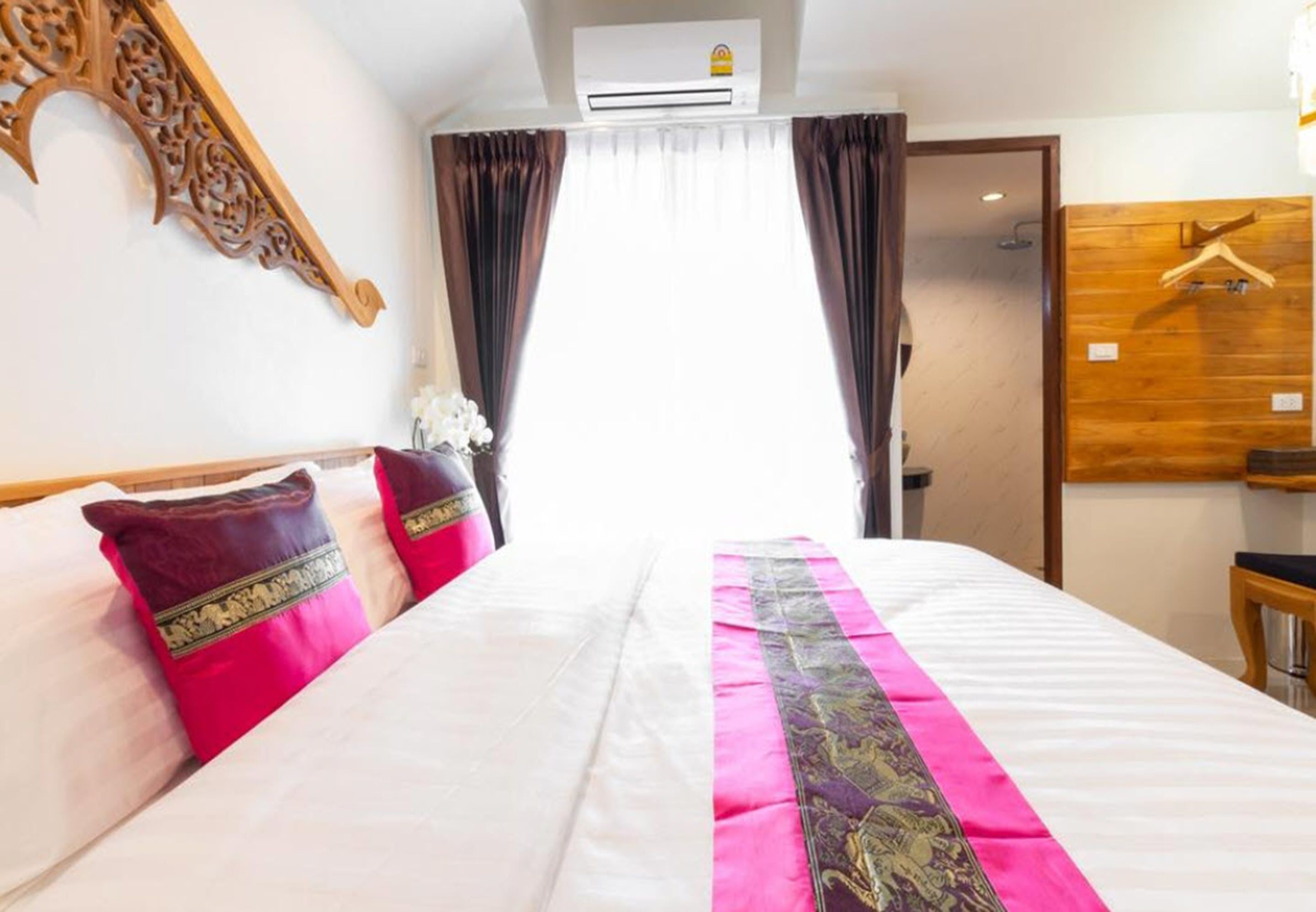 Une chambre d’hôtel avec un grand lit, deux oreillers, une tête de lit décorative, un appareil de climatisation, une armoire en bois et un miroir.