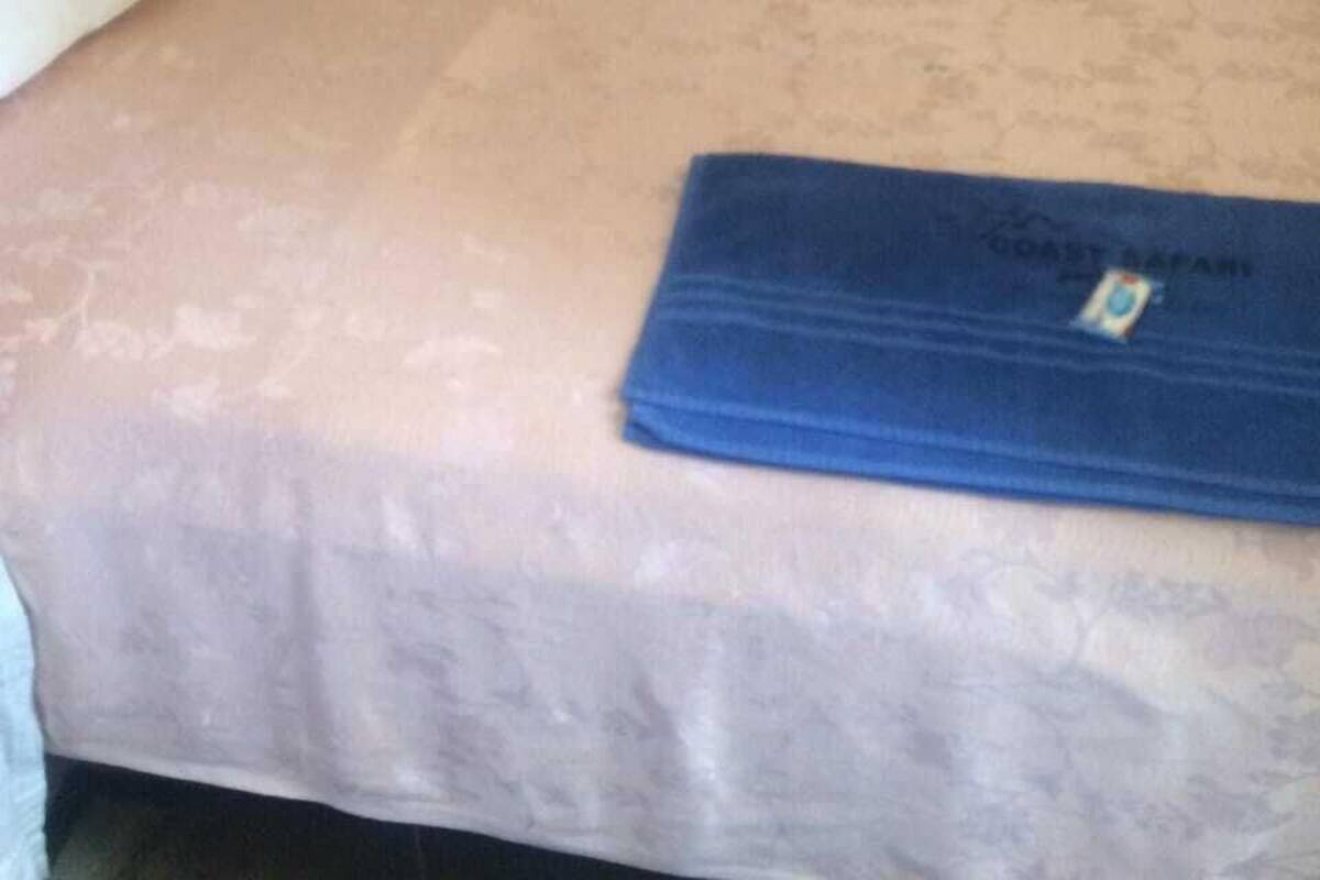A single bed with a pink bedspread and a blue blanket labeled "COAST SAFARI" on a wall with a floral pattern.