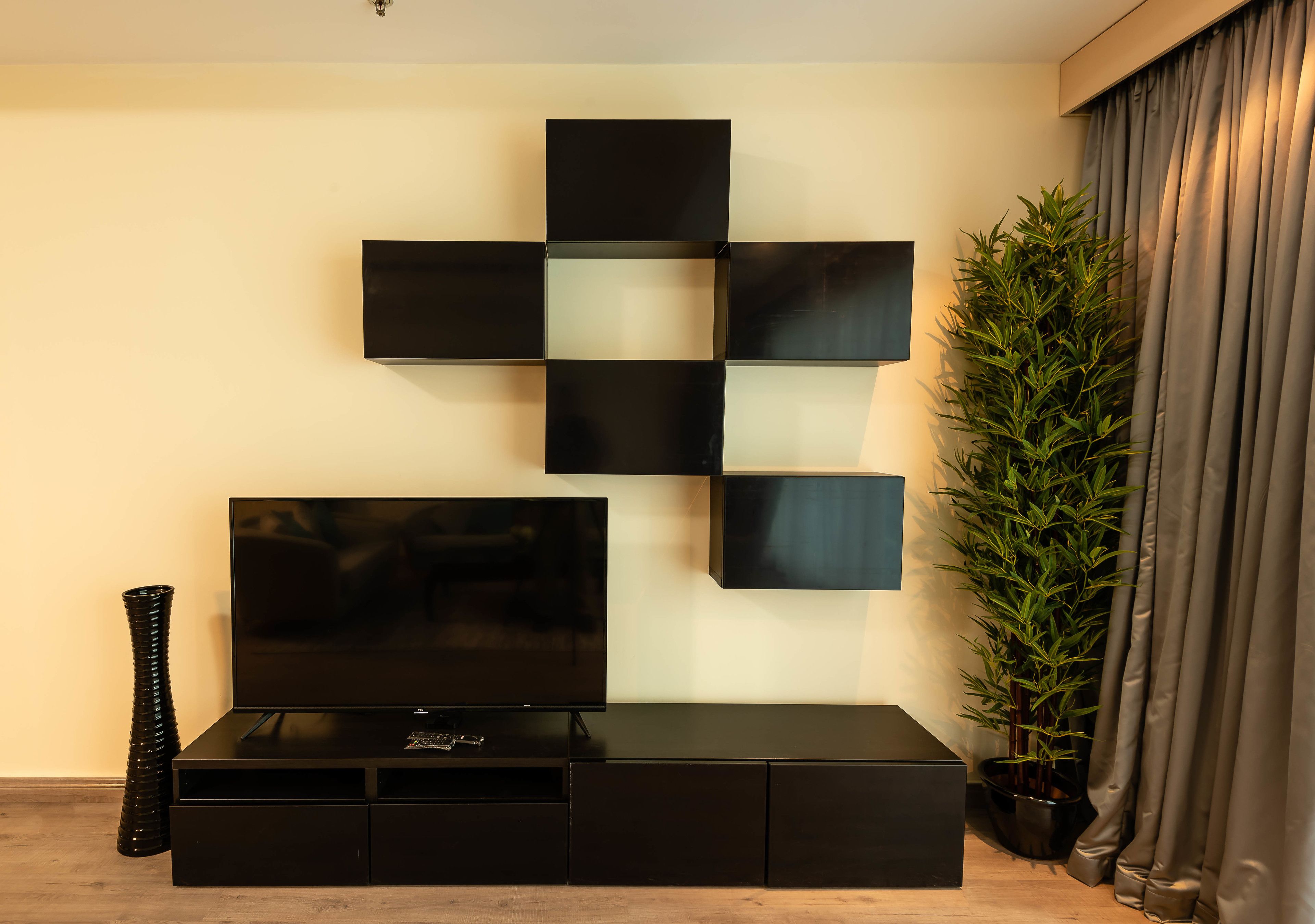 Een moderne woonkamer met een flatscreen televisie op een zwarte tv-kast, een decoratief kunstwerk aan de muur en een hoge groene plant.